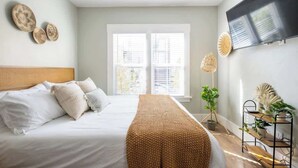 A modern bedroom with a large bed, a TV mounted on the wall, and a window with blinds. - Sunny Stylish & Central - Luxe Little Italy Studio (San Diego)