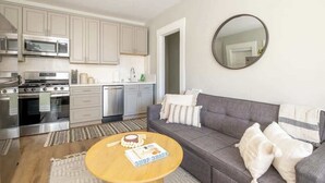 A modern kitchen with stainless steel appliances, a grey sofa, and a round mirror on the wall. - Sunny Stylish & Central - Luxe Little Italy Studio (San Diego)
