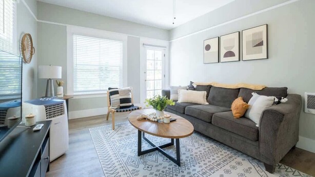 A modern living room with a grey sofa, a coffee table, and a patterned rug. There is a window with blinds, a ceiling fan, and framed artwork on the wall. - Chic Little Italy Apt | Balcony | Walk Everywhere! (San Diego)