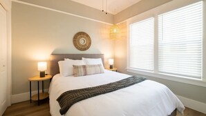 A bedroom with a large bed, two bedside tables with lamps, a wall-mounted circular decoration, and a window with blinds. - Chic Little Italy Apt | Balcony | Walk Everywhere! (San Diego)