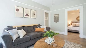 A modern living room with a grey sofa, a round wooden coffee table, and a patterned rug. There are framed artworks on the wall and a doorway leading to a bedroom. - Chic Little Italy Apt | Balcony | Walk Everywhere! (San Diego)