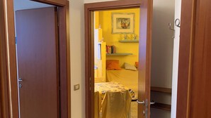 Un couloir étroit avec une porte en bois sur la gauche, qui donne sur une pièce avec un mur jaune, un lit et une petite table.