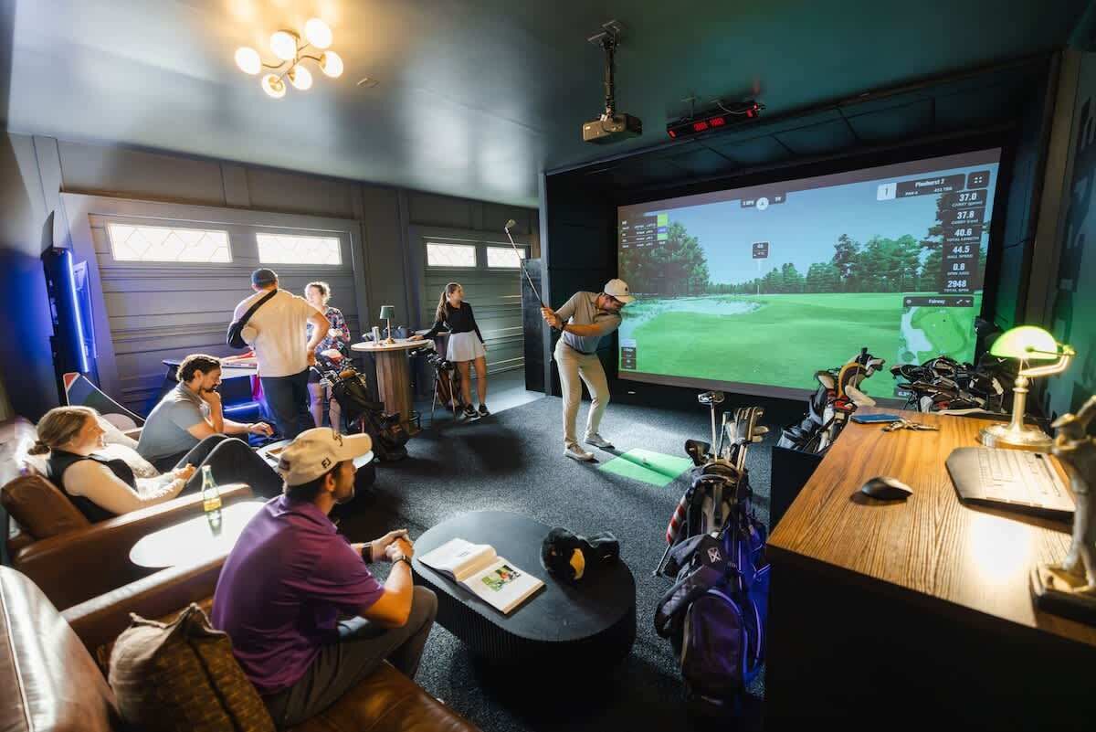 Ett rum för golfsimulering med en stor skärm, golfklubbor och personer som spelar golf.