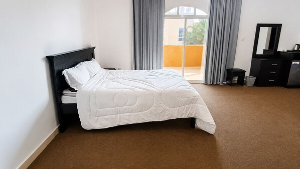 A neatly made bed with a white comforter in a room with a brown carpet, a black nightstand, a black dresser, and a large window with curtains.