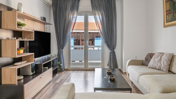 Ein Wohnzimmer mit Fernseher, Holzböden, einer Couch, einem Couchtisch und einem Balkon mit Vorhängen.