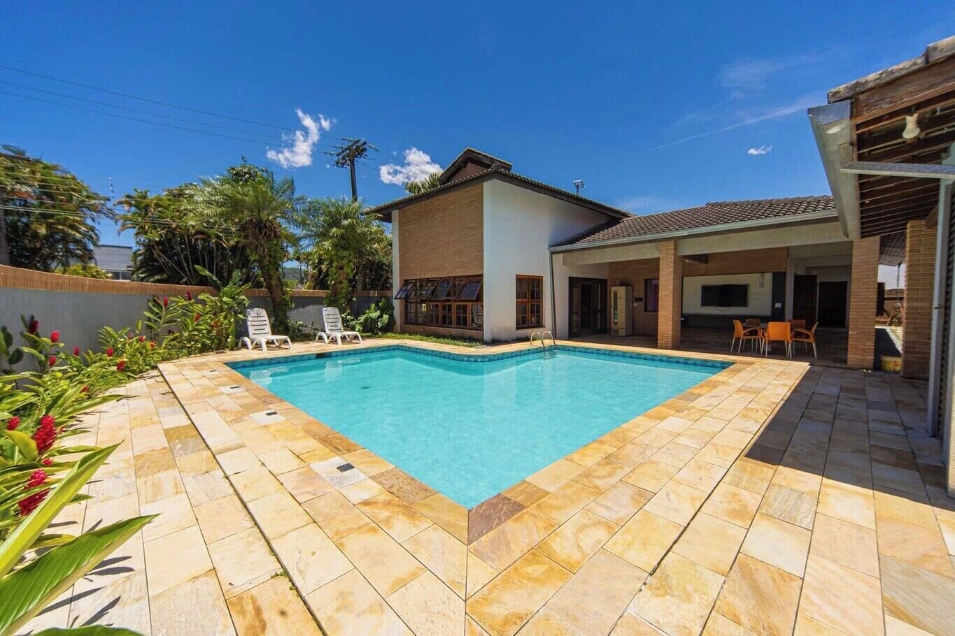 Une piscine rectangulaire, bordée d’une terrasse carrelée, est entourée d’un bâtiment blanc doté d’un toit plat et d’un patio couvert avec des chaises.