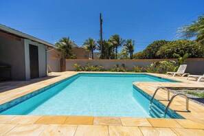A rectangular swimming pool with a tiled deck, surrounded by palm trees and a low wall. - Casa c Piscina e Churrasqueira em Condom nio (Guarujá)