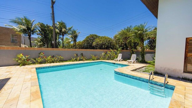 A rectangular swimming pool with a tiled deck, surrounded by palm trees and a low wall. - Casa c Piscina e Churrasqueira em Condom nio (Guarujá)