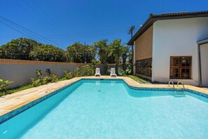A rectangular swimming pool with two white lounge chairs, surrounded by a tiled deck and a low brick wall. - Casa c Piscina e Churrasqueira em Condom nio (Guarujá)