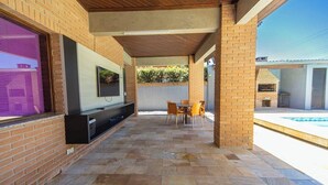 A covered patio with a TV, outdoor seating, and a pool. - Casa c Piscina e Churrasqueira em Condom nio (Guarujá)