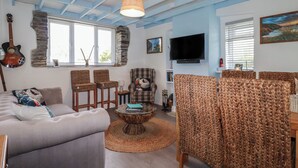 Un salon comprenant un canapé, des fauteuils, une table basse, une guitare, un téléviseur et un mur en pierre.