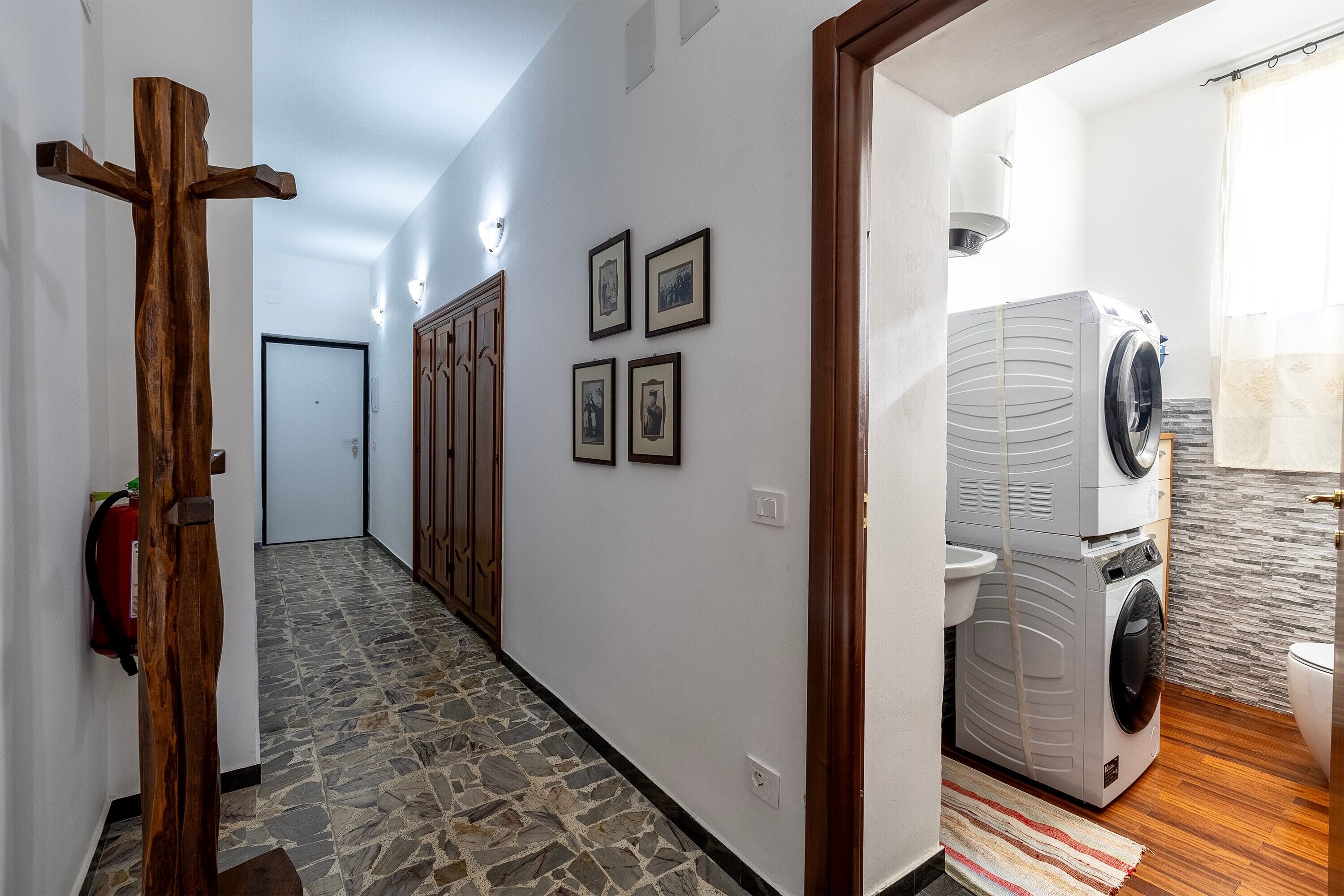 A hallway with a wooden cross, stone flooring, a washing machine, and a toilet in a bathroom.