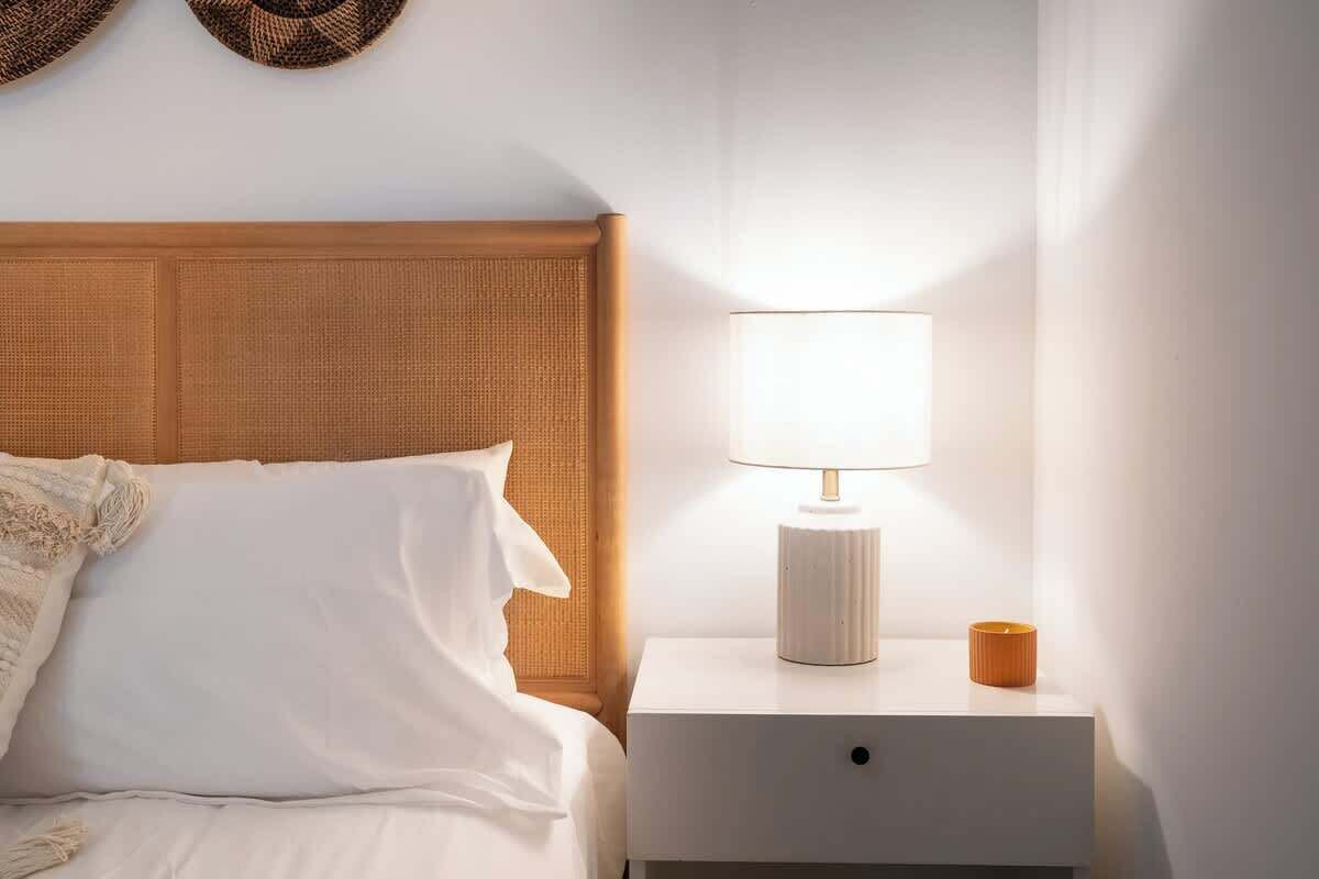 A neatly made bed with white linens and a textured headboard, a bedside table with a lamp and a small orange container, and a wall-mounted basket above the bed.
