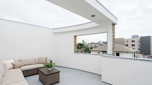Terraço no telhado com um sofá seccional bege, uma mesa de centro de vime e um vaso com planta.