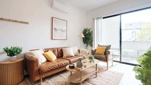 A modern living room with a brown leather sofa, a wooden coffee table, and a large window offering a view of the outdoors. - Modern Central Gem w/ Patio | Steps to All! (San Diego)