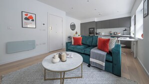A modern living room with a teal sofa, a round coffee table, and a kitchen in the background. - Chic 1BR Close to Tower & Thames (London)