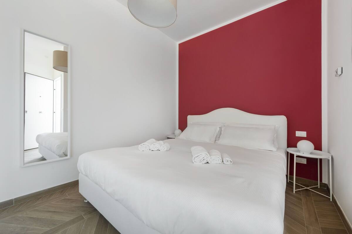 Una habitació d'hotel amb llit, una tauleta de nit, un mirall i una paret vermella decorativa.