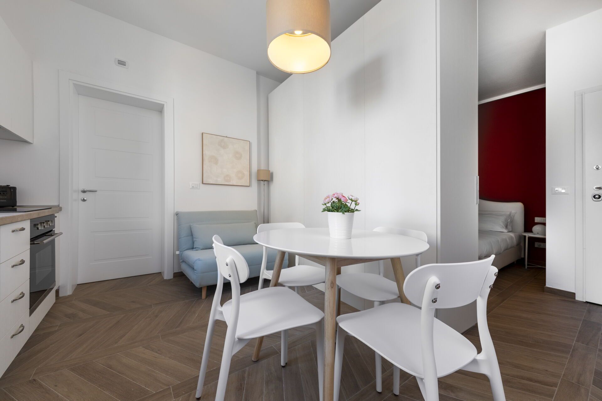 A modern kitchen with white cabinets, a dining table with four chairs, a blue sofa, and a red accent wall.