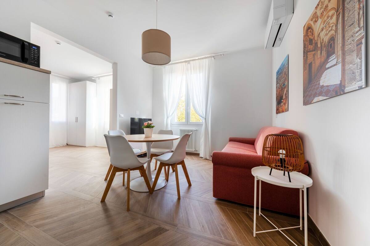 Un espai de vida modern amb zona de menjador, un sofà vermell, una taula petita amb una làmpada i obra d'art a la paret.