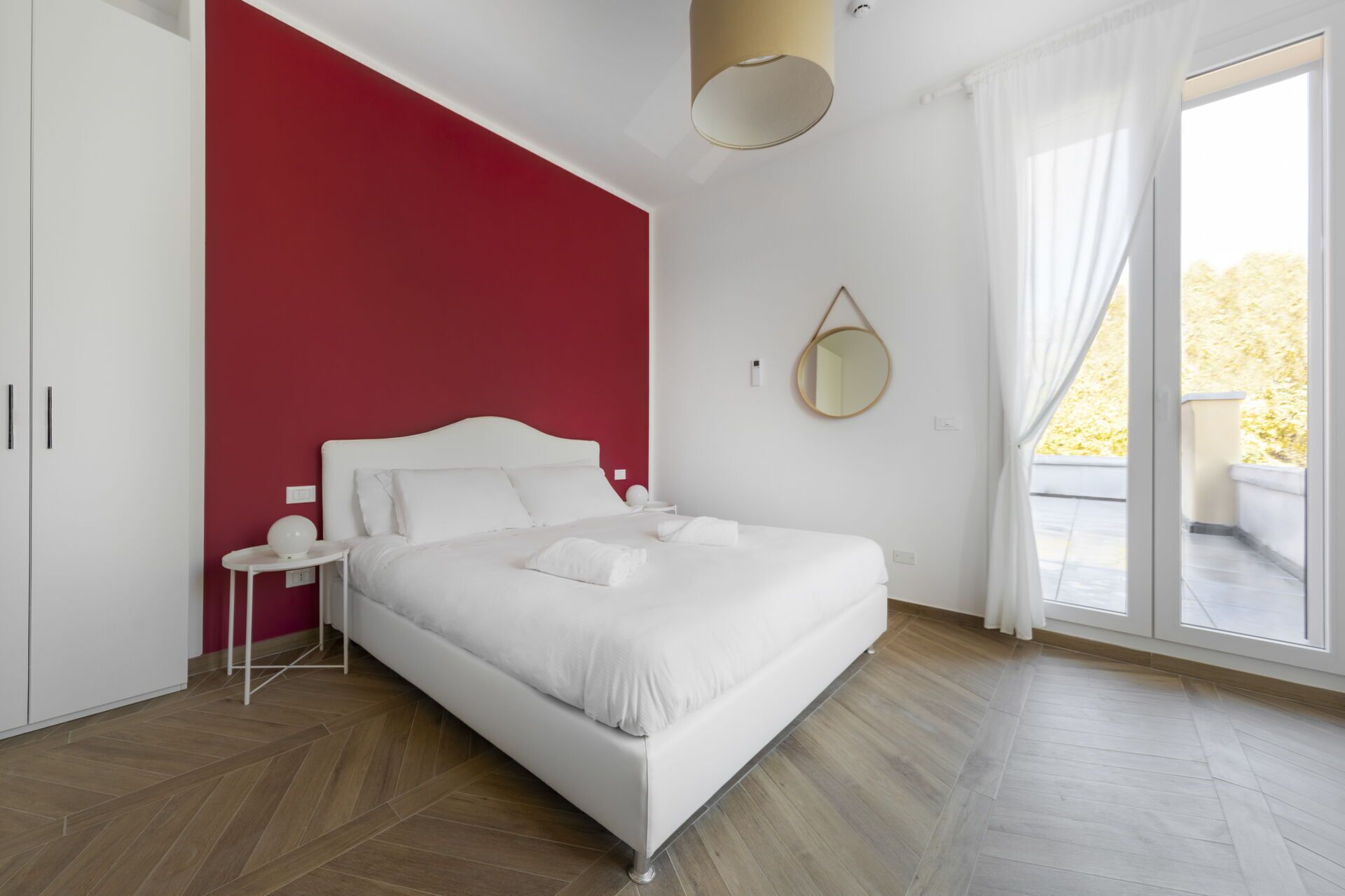 A bedroom with a bed, white nightstand, a red accent wall, a round mirror, and a sliding glass door leading to an outdoor area.