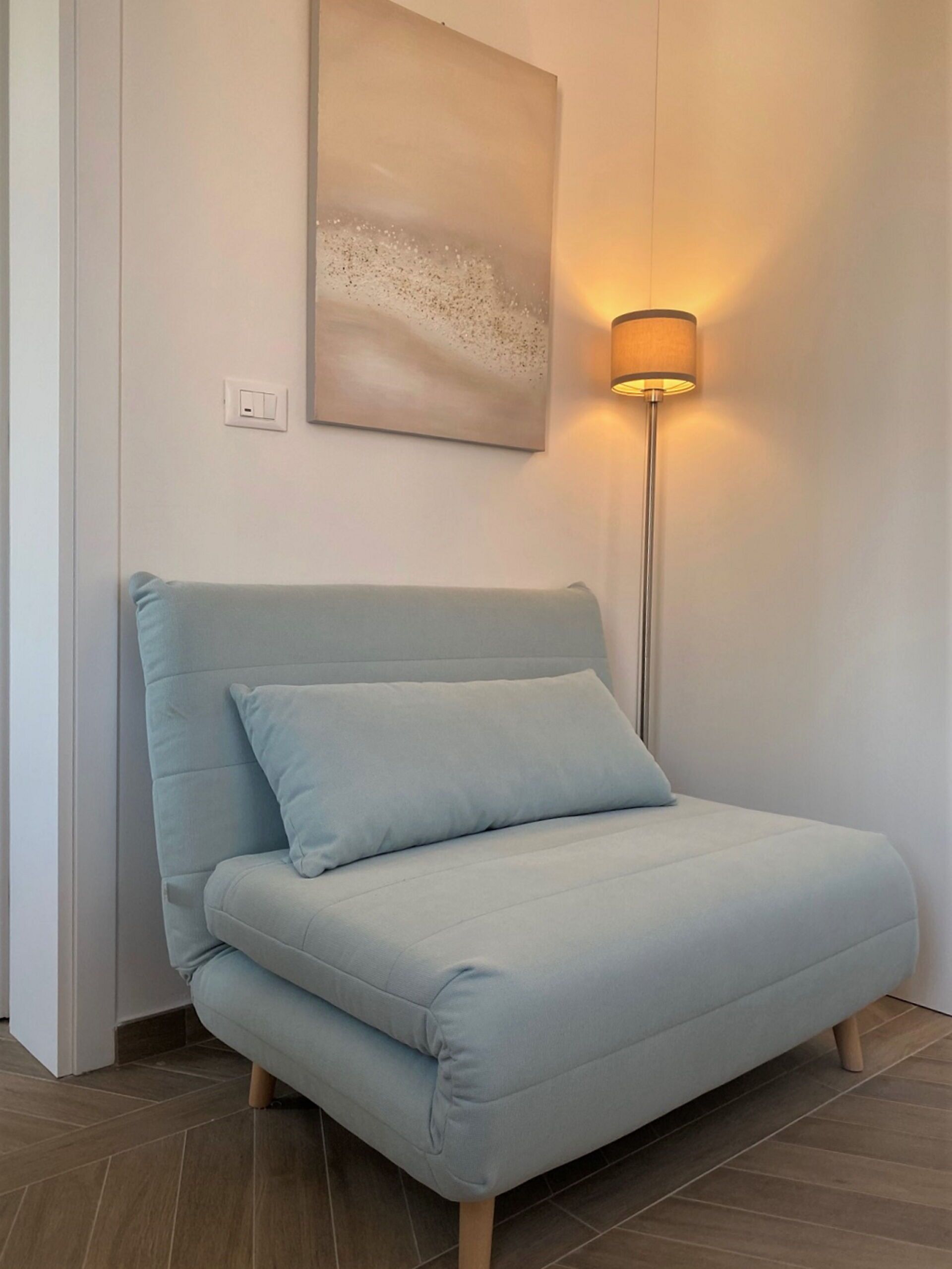 Un sofá azul claro con un cojín, una lámpara de pie y un cuadro abstracto colgado en la pared en una habitación con suelo de baldosas.
