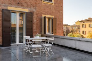 Un balcón con mesa y sillas, un edificio de ladrillo y una ventana con contraventanas.