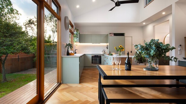 Une cuisine moderne avec une table à manger en bois, des appareils électroménagers intégrés et de grandes fenêtres donnant sur un jardin.