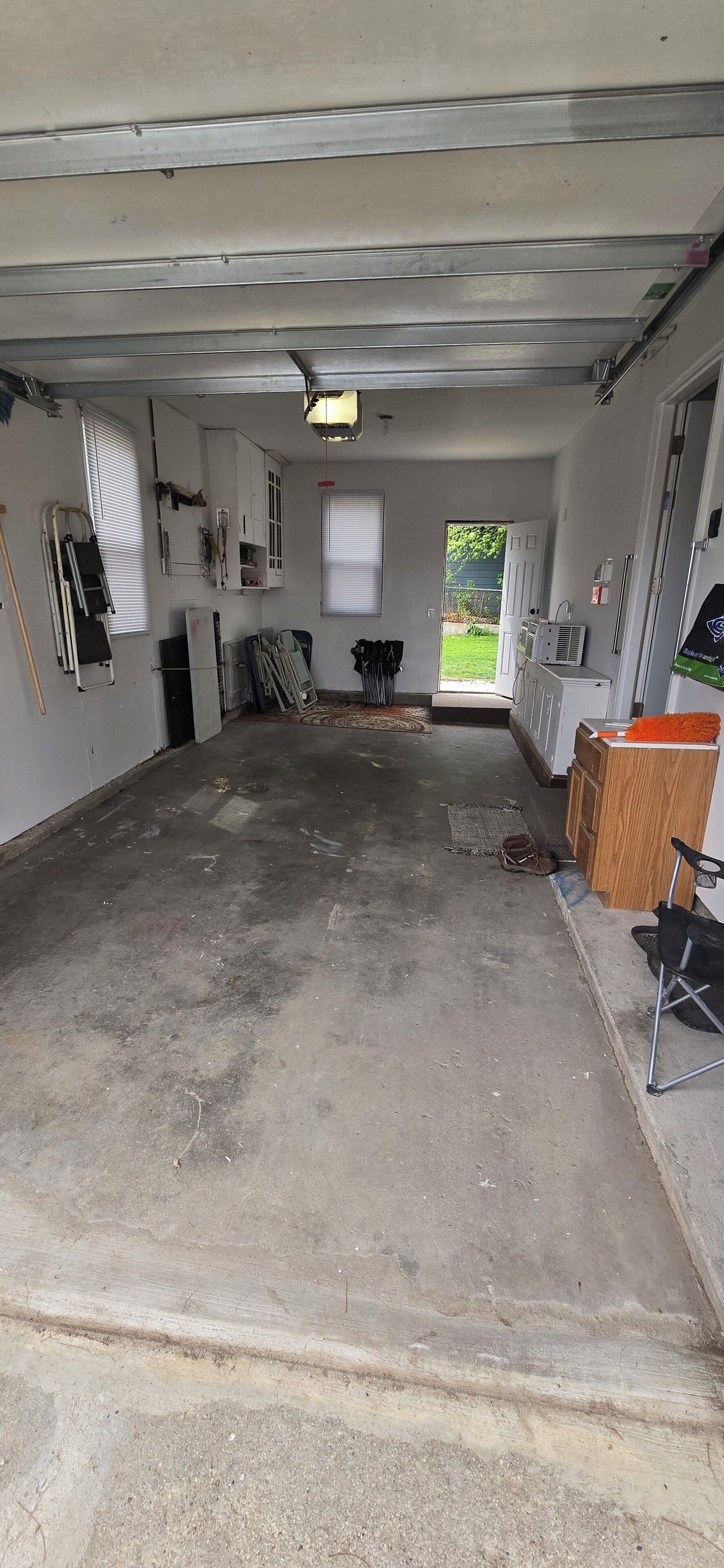 A garage with a concrete floor, a door leading to a backyard, and various items like a ladder, a radiator, and a folding chair.
