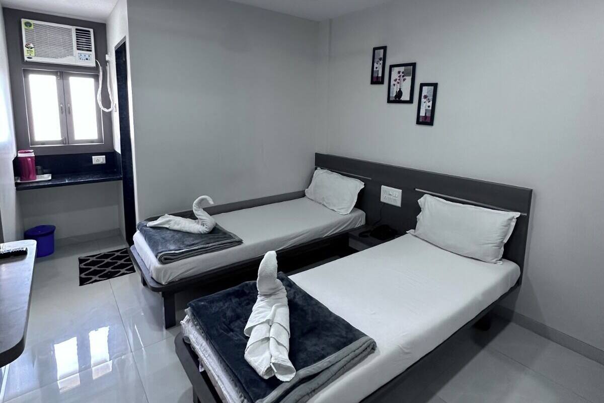 A hotel room with two single beds, a small desk, a window with an air conditioning unit, and a ceiling light.