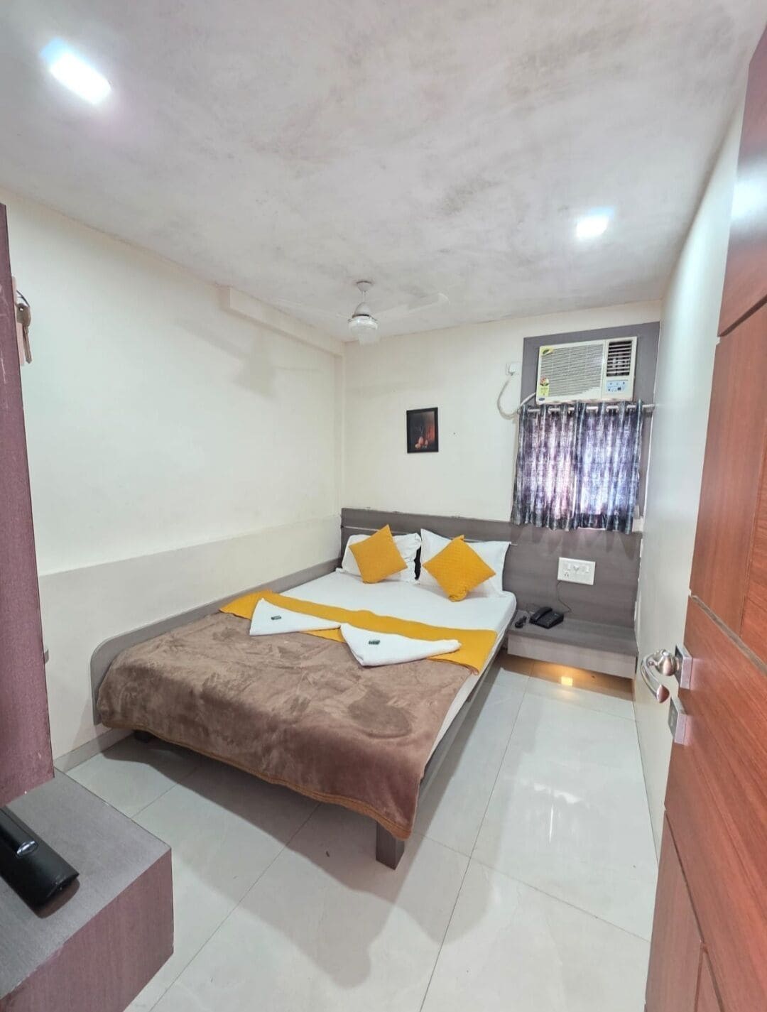A hotel room with a bed, a bedside table, a wall-mounted air conditioner, and a ceiling fan.