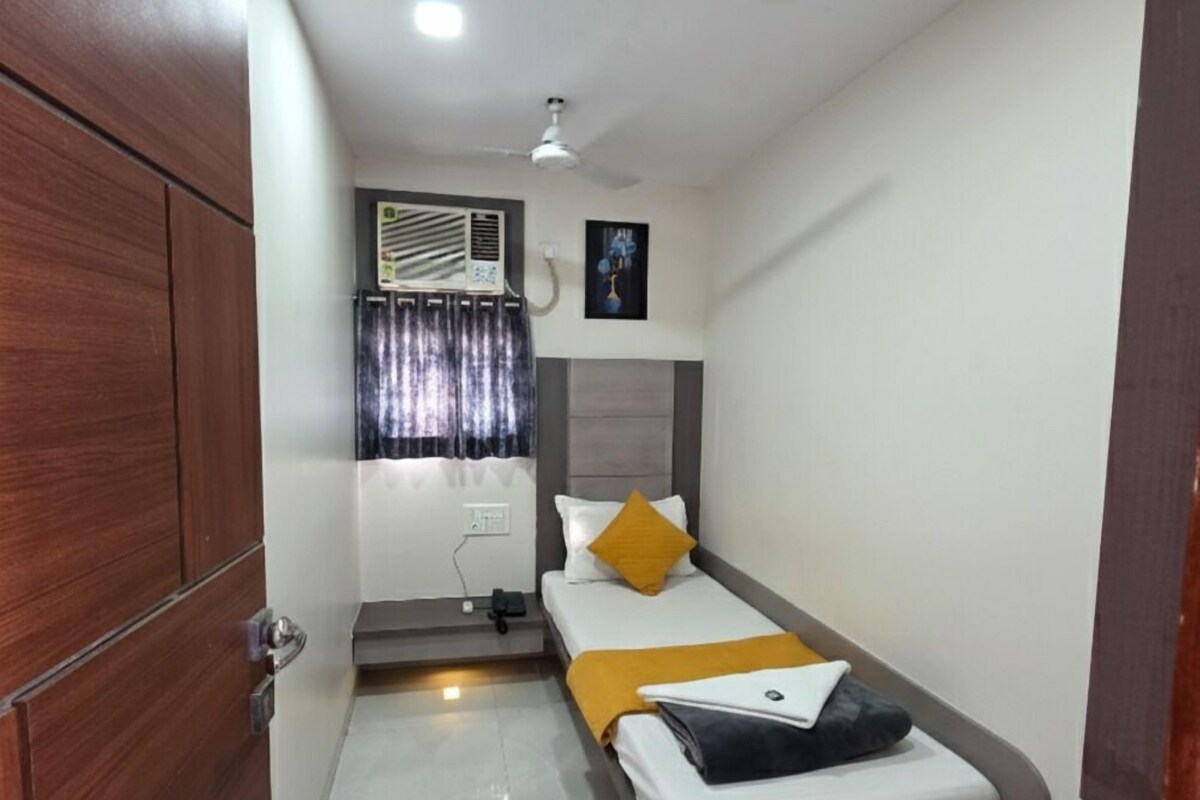 Une chambre d’hôtel de taille réduite, comprenant un lit simple, une petite table de chevet, une armoire, un ventilateur de plafond et une fenêtre avec des rideaux.