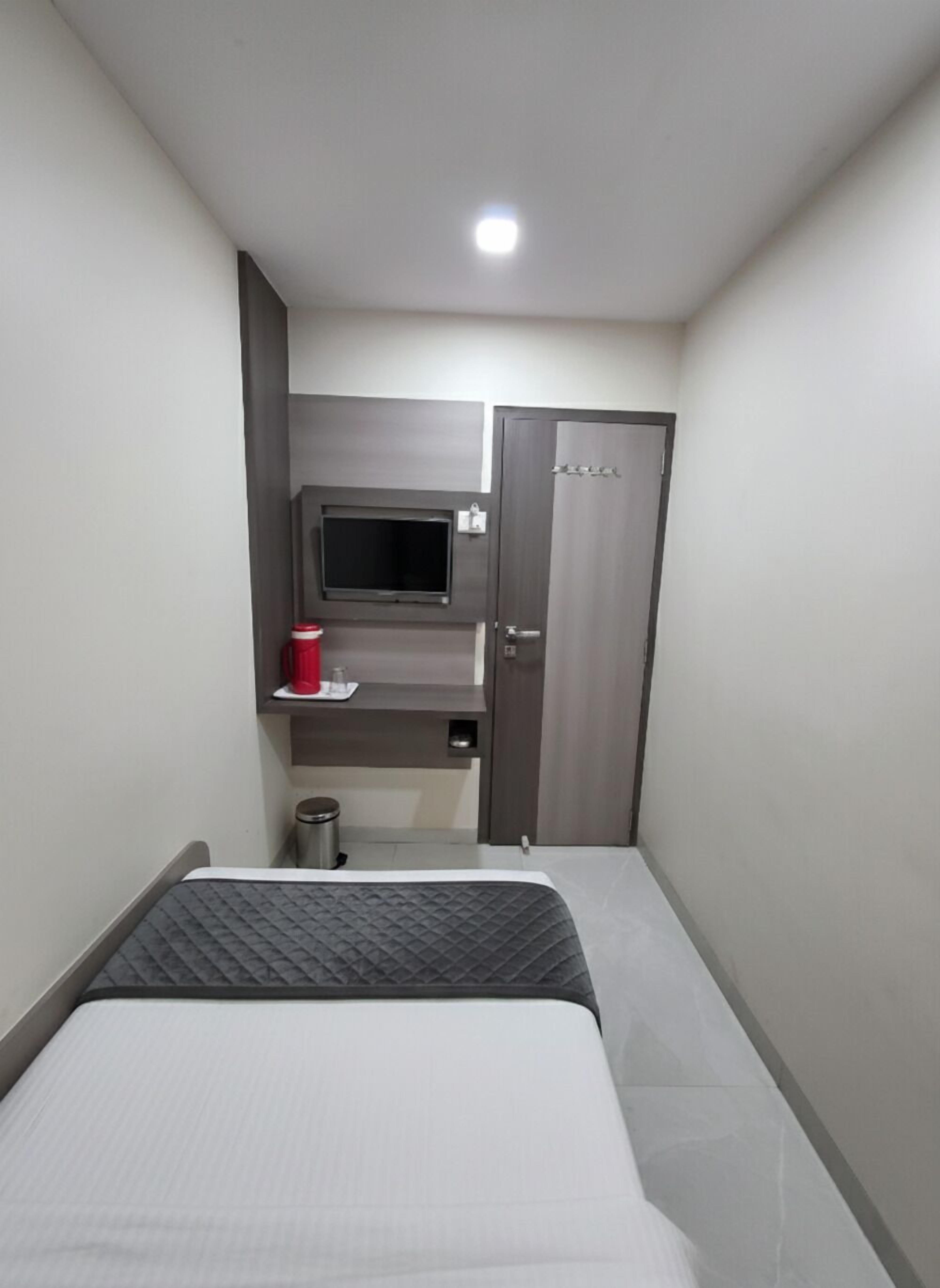 Une petite chambre d’hôtel moderne, équipée d’un lit, d’un téléviseur fixé au mur, d’une armoire et d’une petite étagère sur laquelle se trouve un récipient rouge.