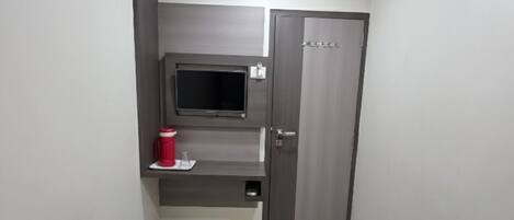 A small, modern hotel room with a bed, a TV mounted on the wall, a wardrobe, and a small shelf with a red container.
