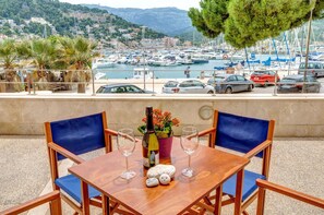 Ein Holztisch mit zwei blauen Stühlen, einer Weinflasche und zwei Weingläsern auf einer Terrasse mit Blick auf einen Yachthafen und eine Küstenstadt.