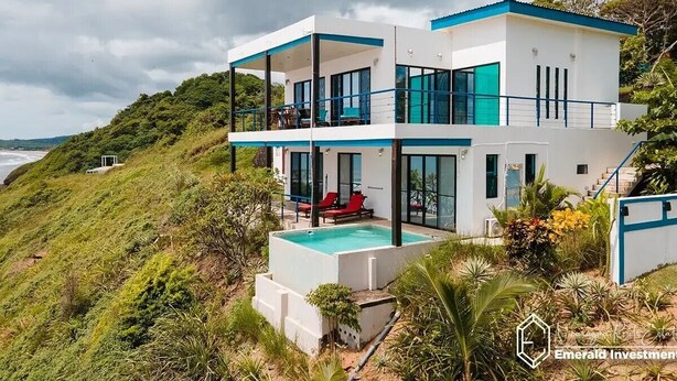 Una moderna casa a due piani con piscina, situata su una collina con vista sull'oceano.