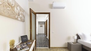 Et hotelværelse med seng, et natbord, en lampe, et skrivebord med en computer, en vægmonteret klimaanlæg og en gang med flisegulv samt en dør, der fører til et andet rum.