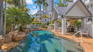 A swimming pool with a rocky edge, surrounded by palm trees and a covered patio area with outdoor seating. - The Bay Apartments 15 - Rainbow Bay - Linen Provided - Min. 2 Night Stays Year Round (Coolangatta)