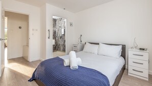 A bedroom with a large bed, a bedside table, a nightstand, and a bathroom visible through an open door. - Baird Place (No 26) | Elie (Elie)