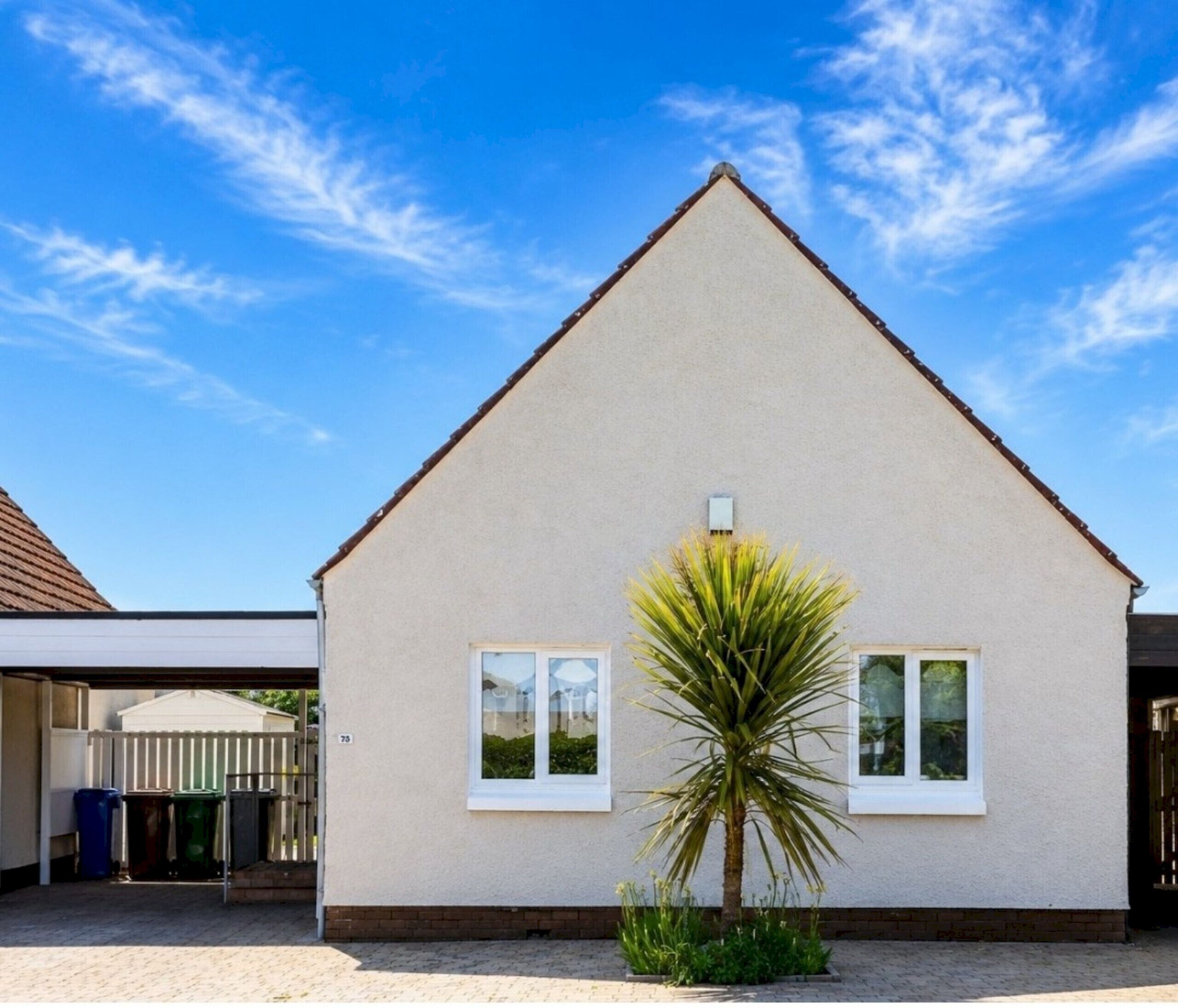 A single-story house with a triangular roof, a small window, and a palm tree in front.
