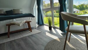 Ein Schlafzimmer mit einem Bett, einer Bank, einem Sessel, einer Topfpflanze und Blick auf einen Garten durch große Fenster.