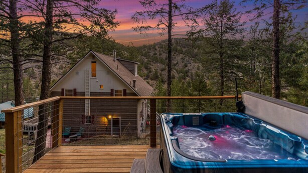 A wooden deck with a hot tub, a house, and a forested backdrop.
