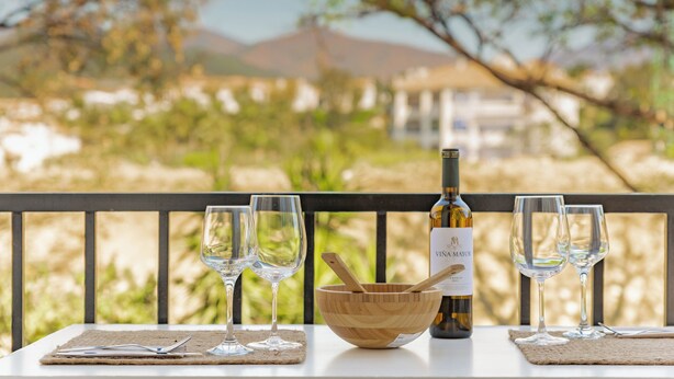 Una mesa con copas de vino, una botella y un cuenco de madera en un balcón con vistas a un paisaje pintoresco.