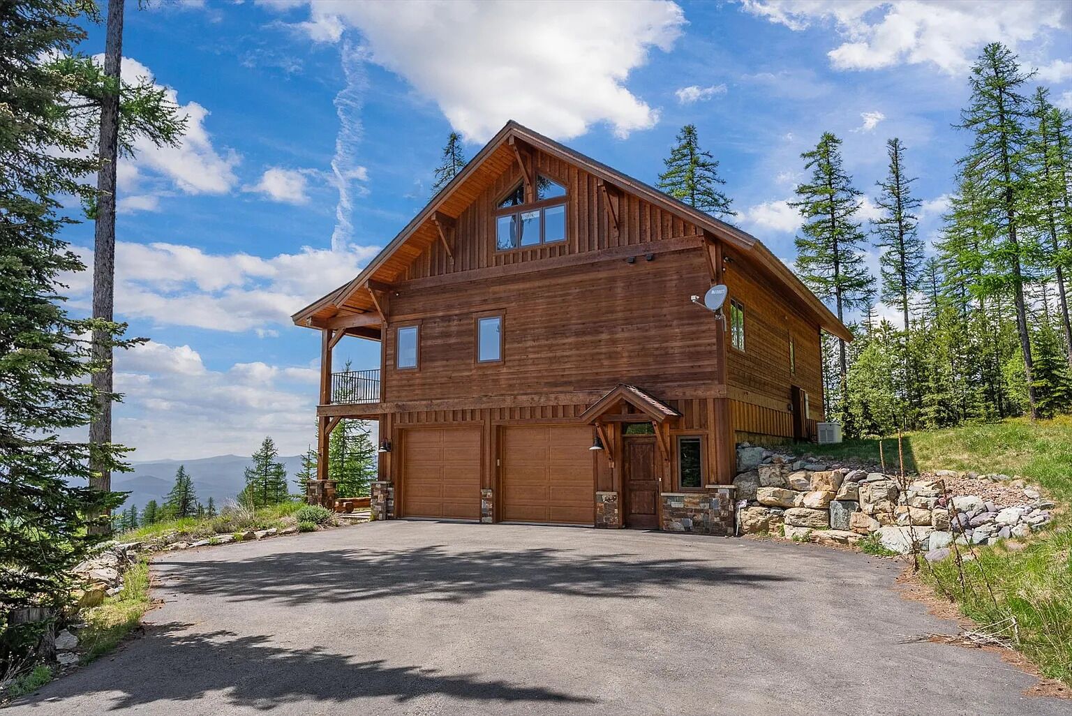 A two-story wooden cabin with a garage, surrounded by trees and a clear sky.