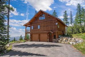 Eine zweistöckige Holzhütte mit Garage, umgeben von Bäumen und blauem Himmel.
