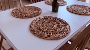 A dining table with woven placemats, a vase with dried flowers, and a purple sofa in the background.