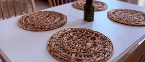 A dining table with woven placemats, a vase with dried flowers, and a purple sofa in the background.