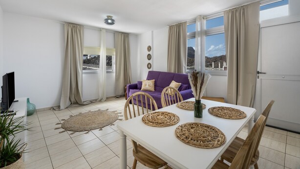 A living room with a white table, beige curtains, a purple sofa, and a view of the outdoors.