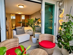 A modern living room with a sofa, a wicker chair with a red cushion, and a potted plant. - Serene Coastal Studio with King Bed & Balcony (Luquillo)