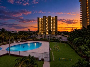 A swimming pool with a fenced area, palm trees, and a large building with multiple floors in the background. - Serene Coastal Studio with King Bed & Balcony (Luquillo)