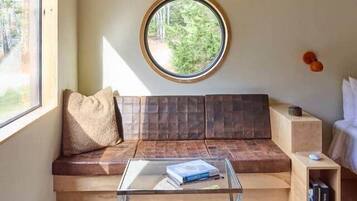 Ein gemütliches Zimmer mit Holzbalkendecke, einem Rundfenster, einer Couch, einem Glastisch und einem kleinen Bücherregal.